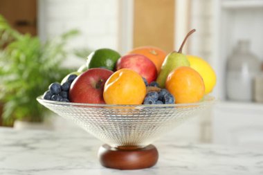 Beyaz mermer masanın üzerinde farklı taze meyveler bulunan cam vazo, yakın plan.