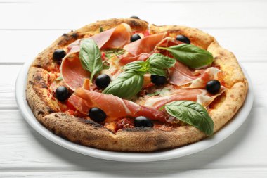 Pişmiş jambonlu lezzetli pizza, zeytin, güneşte kurutulmuş domates ve beyaz ahşap masada fesleğen, yakın plan.
