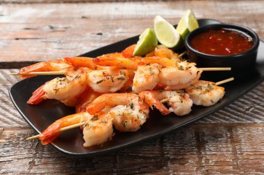 Ahşap masada sos ve limonla servis edilen leziz ızgara karides şişleri.