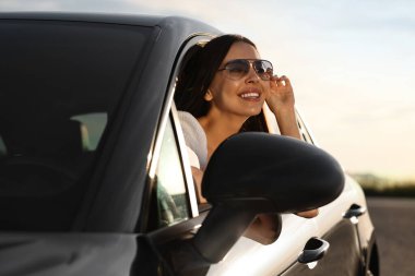 Dışarıdaki arabanın camından sarkan güneş gözlüklü, gülümseyen genç kadın. İyi yolculuklar.