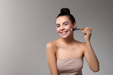 Gülümseyen kadın açık gri arka planda fırçayla makyaj yapıyor. Metin için boşluk