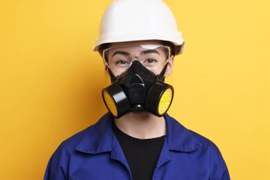 Solunum cihazı, koruyucu gözlük ve turuncu arka planda kask takan bir işçi.