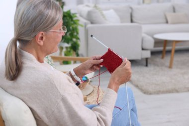 Örgü iğneli yaşlı kadın evde desenlere bakıyor.