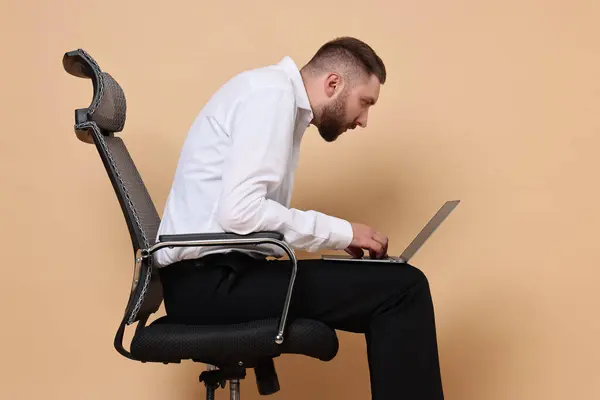 stock image Man with poor posture sitting on chair and using laptop against pale orange background