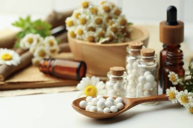 Homeopatik tedavi. Haplı kaşık, şişeler ve papatya çiçekleri beyaz masada, yakın plan.