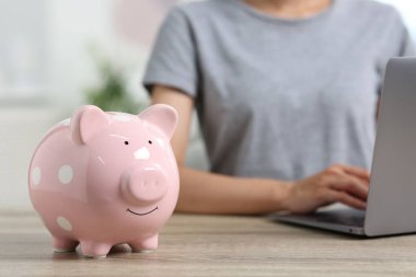 Woman using laptop at wooden table, focus on piggy bank indoors clipart