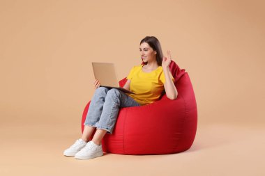 Bilgisayarlı gülümseyen kadın bej arka planda kırmızı fasulye koltukta otururken online toplantı yapıyor.