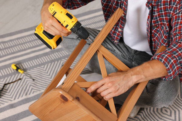 Man repairing wooden stool with electric screwdriver indoors, closeup
