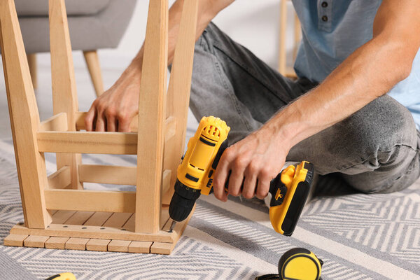 Man repairing wooden stool with electric screwdriver indoors, closeup
