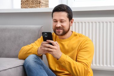 Evdeki kaloriferin yanında akıllı telefon kullanan mutlu bir adam.