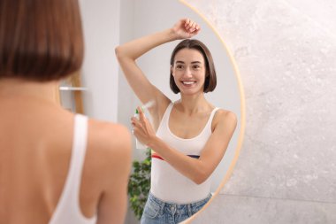 Gülümseyen kadın evde aynanın yanında sprey deodorant sürüyor.