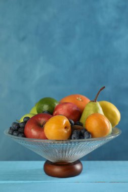 Açık mavi ahşap masada farklı taze meyveler bulunan cam vazo, metin için yer.