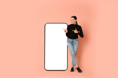 Açık mercan arka planında boş ekranı olan büyük bir cep telefonunu işaret eden güzel genç bir kadın.