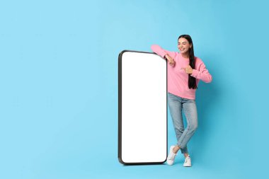 Güzel genç bir kadın açık mavi arka planda boş ekranlı büyük bir cep telefonunu işaret ediyor.