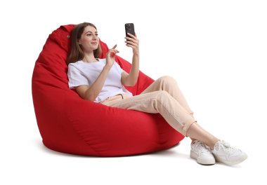 Akıllı telefonlu güzel genç bir kadın beyaz arka planda kırmızı fasulye koltukta otururken online toplantı yapıyor.