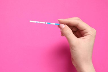 Kadının elinde pembe arka planda hamilelik testi var.
