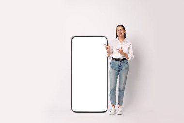 Güzel genç bir kadın arka planında boş ekran olan büyük bir cep telefonunu işaret ediyor.