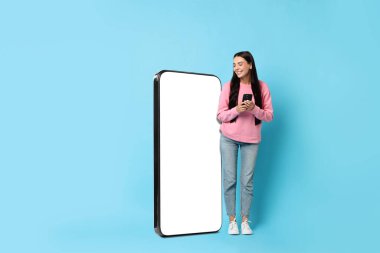 Akıllı telefonlu güzel bir kadın açık mavi arka planda boş ekranlı büyük bir cep telefonunun yanında duruyor.