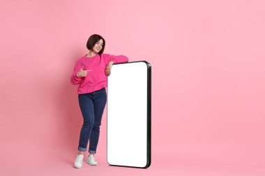 Pembe arka planda boş ekranı olan büyük cep telefonunu gösteren güzel genç bir kadın.