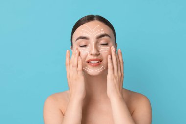 Güzel genç bir kadın açık mavi arka planda parmaklarıyla yüz masajı yapıyor. Hareket yönlerini gösteren cilt üzerindeki çizgiler