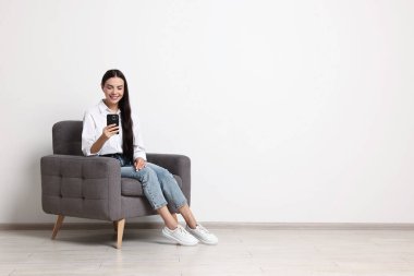 Akıllı telefonlu gülümseyen kadın beyaz duvara karşı koltukta oturuyor. Metin için boşluk