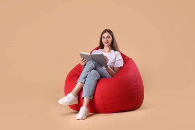 Bej arka planda kırmızı fasülye koltuğu üzerine kitap okuyan güzel genç bir kadın.