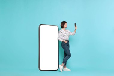 Akıllı telefonlu güzel bir kadın açık mavi arka planda boş ekranlı büyük bir cep telefonunun yanında duruyor.