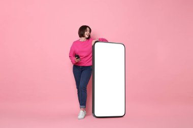 Pembe arka planda boş ekranı olan büyük bir cep telefonunun yanında duran güzel genç bir kadın.