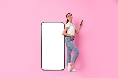 Akıllı telefonlu güzel bir kadın pembe arka planda boş ekranlı büyük bir cep telefonunun yanında duruyor.