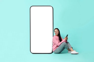 Akıllı telefonlu güzel genç bir kadın turkuaz arka planda boş ekranlı büyük bir cep telefonunun yanında oturuyor.