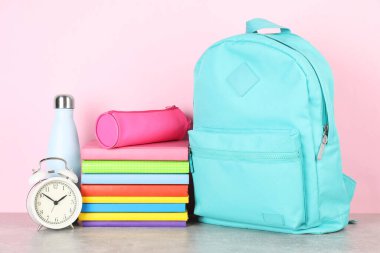 Sırt çantası farklı okul kırtasiyesi, çalar saat ve şişe açık gri masa üzerinde pembe arka plan