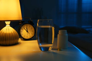 Insomnia treatment. Glass of water, pills and alarm clock on bedside table in bedroom at night clipart