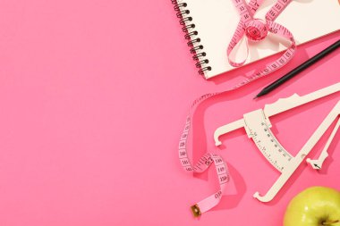 Vücut yağlı kalibre, mezura, pembe arka planda defter ve elma, düz yatış. Metin için boşluk