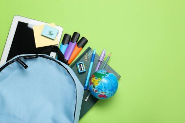 Açık mavi okul çantası, yeşil arka planda kırtasiye malzemesi, üst manzara. Metin için boşluk