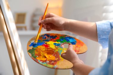 Kadın palette boyayla fırçayı birbirine karıştırıyor, kapalı alanda, yakın planda.