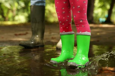 Anne ve kızı lastik botlar giyip açık havada su birikintisinde dikiliyorlar.