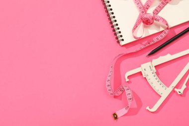Vücut yağ kalibreli, mezura, pembe arka planda defter ve kalem, düz konum. Metin için boşluk