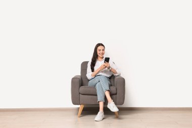 Akıllı telefonlu gülümseyen kadın beyaz duvara karşı koltukta oturuyor.