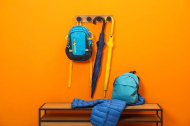 Turuncu duvarın yanındaki ahşap bankta renkli şemsiyeler, sırt çantaları ve ceket