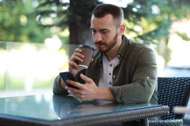 Akıllı telefon kullanıp açık kafede kahve içen yakışıklı bir adam.