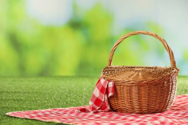 Bir piknik sepeti, kareli peçete ve çimlerin üzerindeki battaniyeyle bulanık yeşil arka plan. Metin için boşluk