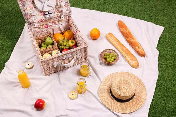 Masa takımlı piknik sepeti, farklı aperatifler, meyve suyu ve açık yeşil çimlerde saman şapka.