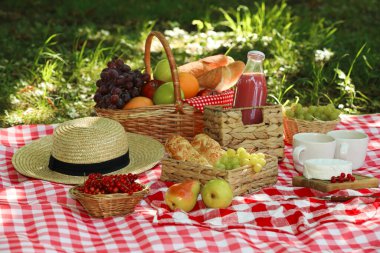 Picnic wicker baskets with delicious food, drink and tableware on checkered blanket outdoors clipart