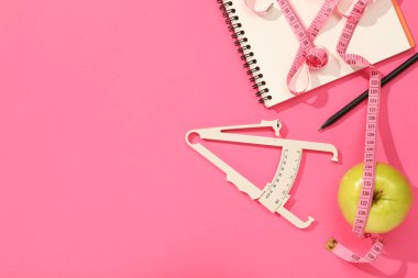 Vücut yağlı kalibre, mezura, pembe arka planda defter ve elma, düz yatış. Metin için boşluk