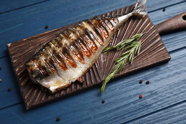 stock image Board with delicious grilled mackerel, peppercorns and rosemary on blue wooden table, above view