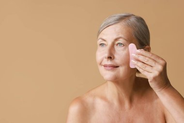 Güzel bir kadın bej arka planda gua sha aracı ile yüz masajı yapıyor, mesaj için boşluk