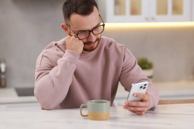 Mutfaktaki beyaz masada akıllı telefon kullanan yakışıklı adam.