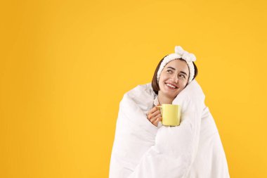Sarı arka planda battaniyeye sarılı fincanı olan mutlu genç bir kadın. Metin için boşluk