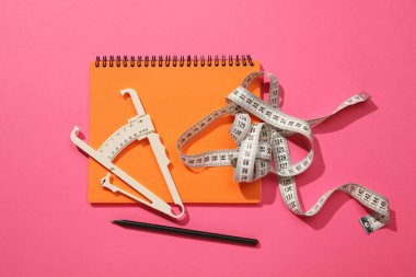 Vücut yağ kalibreli, mezura, pembe arka planda defter ve kalem, düz uzanma