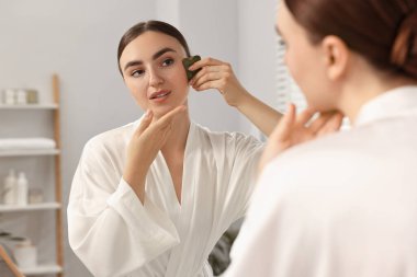 Güzel genç bir kadın evde aynanın yanında gua sha aletiyle yüz masajı yapıyor.